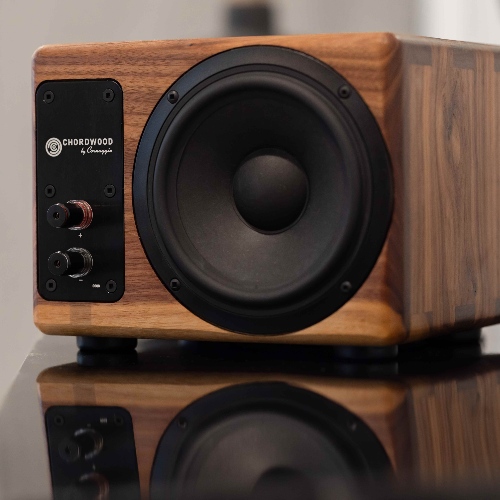 Full view of a single speaker with a walnut wood enclosure, sitting on a reflective surface. It features a large woofer and a black control panel on the left labeled "CHORDWOOD by Cornaggia."