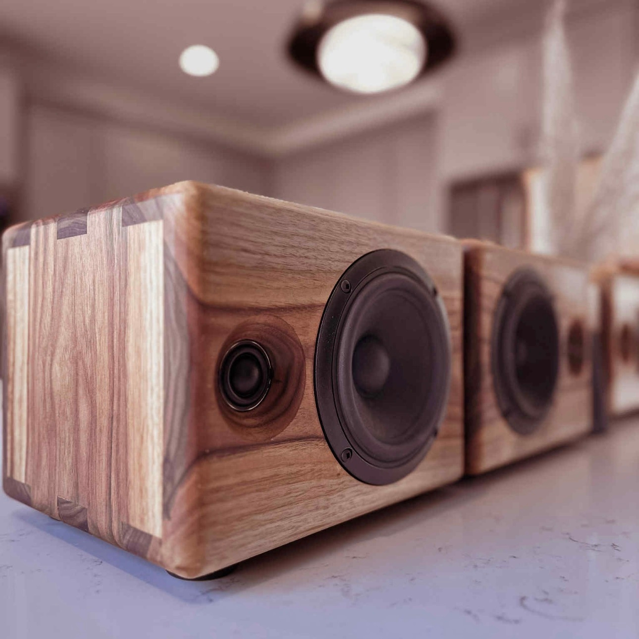 Close-up of a long, horizontal Chordwood by Cornaggia speaker unit crafted from walnut wood. The speaker features visible box joints on the corners, housing multiple black speaker drivers (a tweeter and two woofers visible). The speaker rests on a light kitchen countertop with blurred ceiling lights in the background.