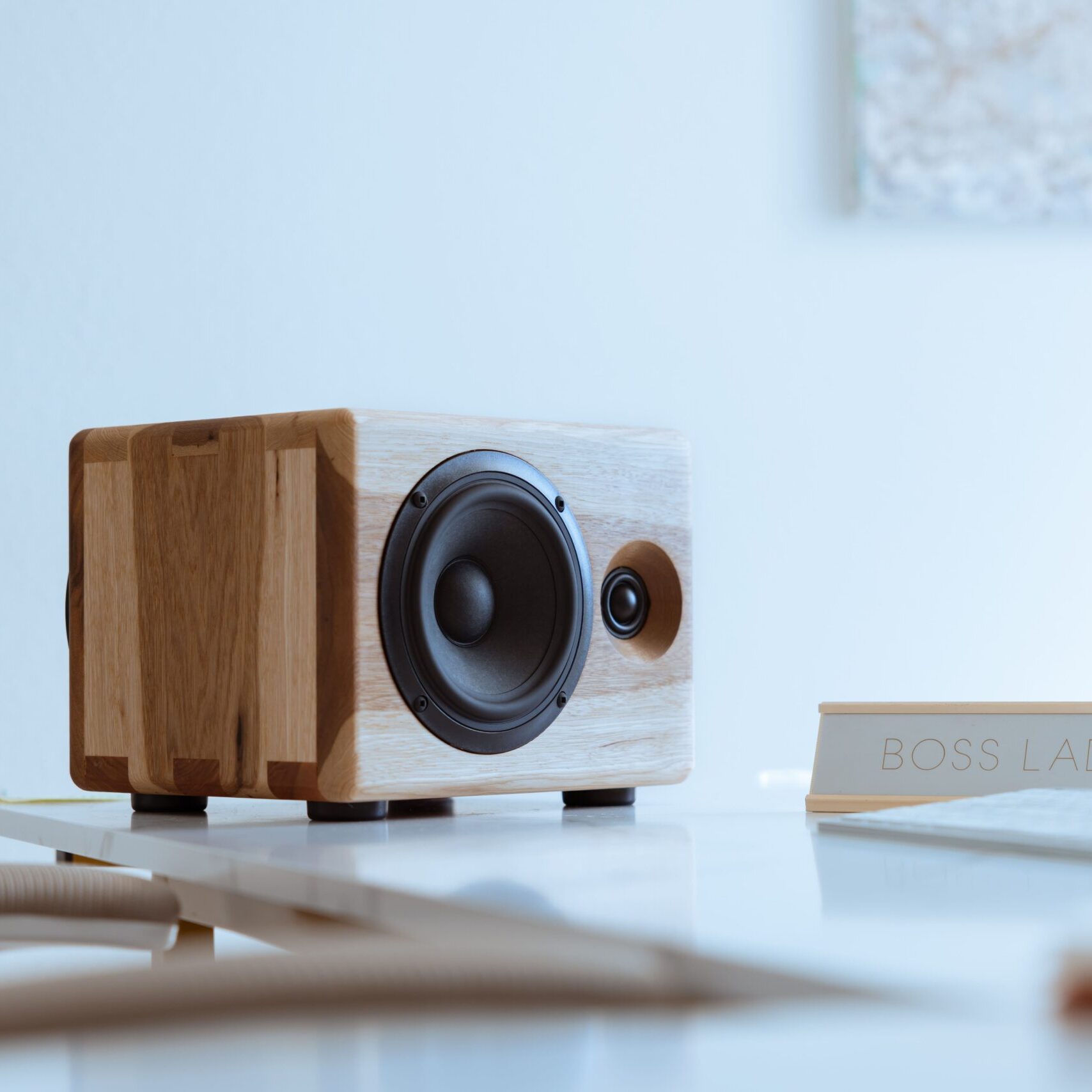 A single Chordwood speaker by Cornaggia, featuring a light-toned wood cabinet with dark contrasting side panels, sits on a bright, minimalist white desk. The speaker's black woofer and tweeter are clearly visible. In the foreground, a small, horizontal sign reads "BOSS LADY," suggesting a professional or home office environment. The room is brightly lit, emphasizing a clean and focused workspace aesthetic.