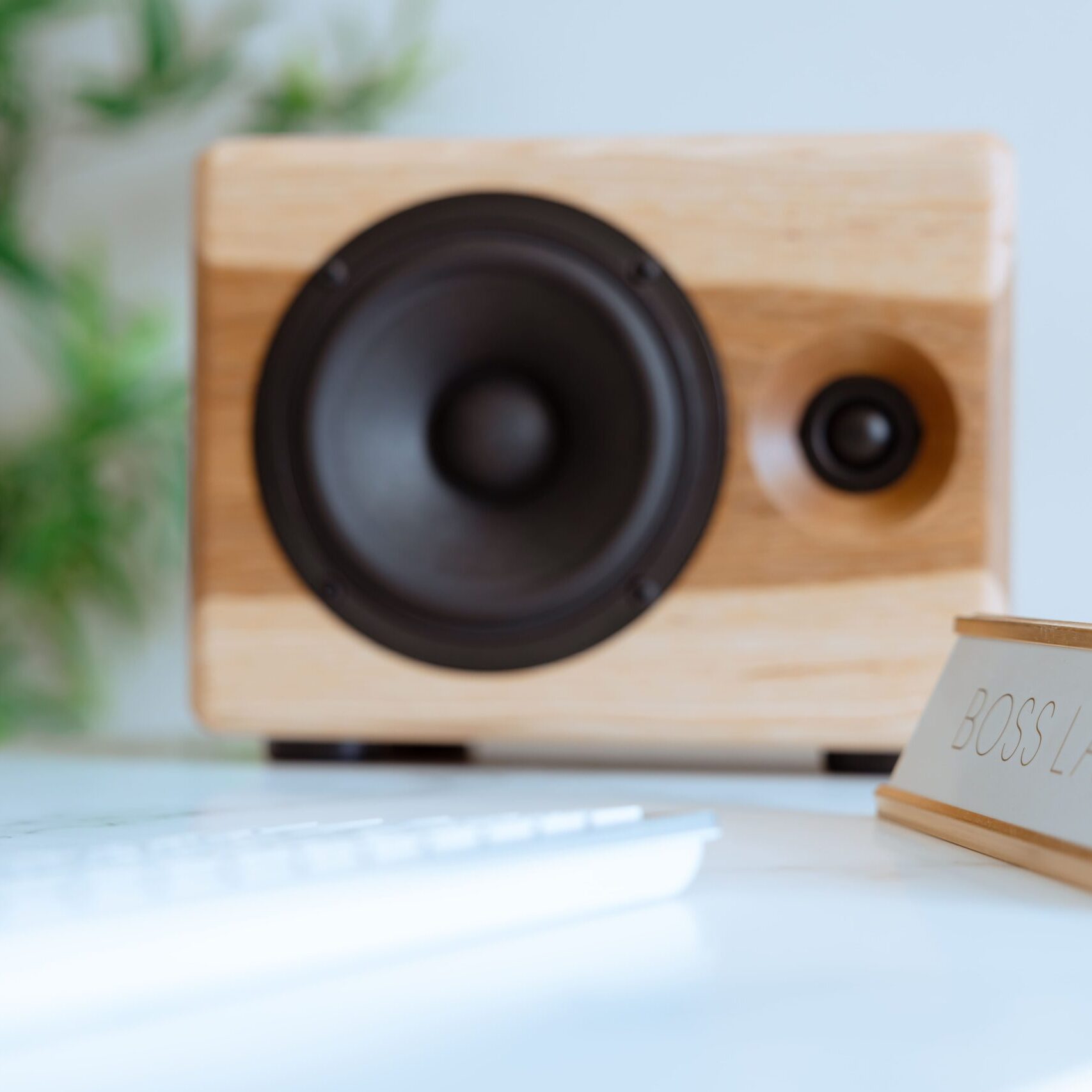 A tight, shallow-depth-of-field close-up of a single Chordwood speaker by Cornaggia, featuring light-toned wood with contrasting vertical side panels, sitting on a white desk. The speaker's black woofer and tweeter are the main focus. In the foreground right, a desk nameplate reading "BOSS LADY" is visible next to a blurred white keyboard, highlighting a professional and stylish home office environment.