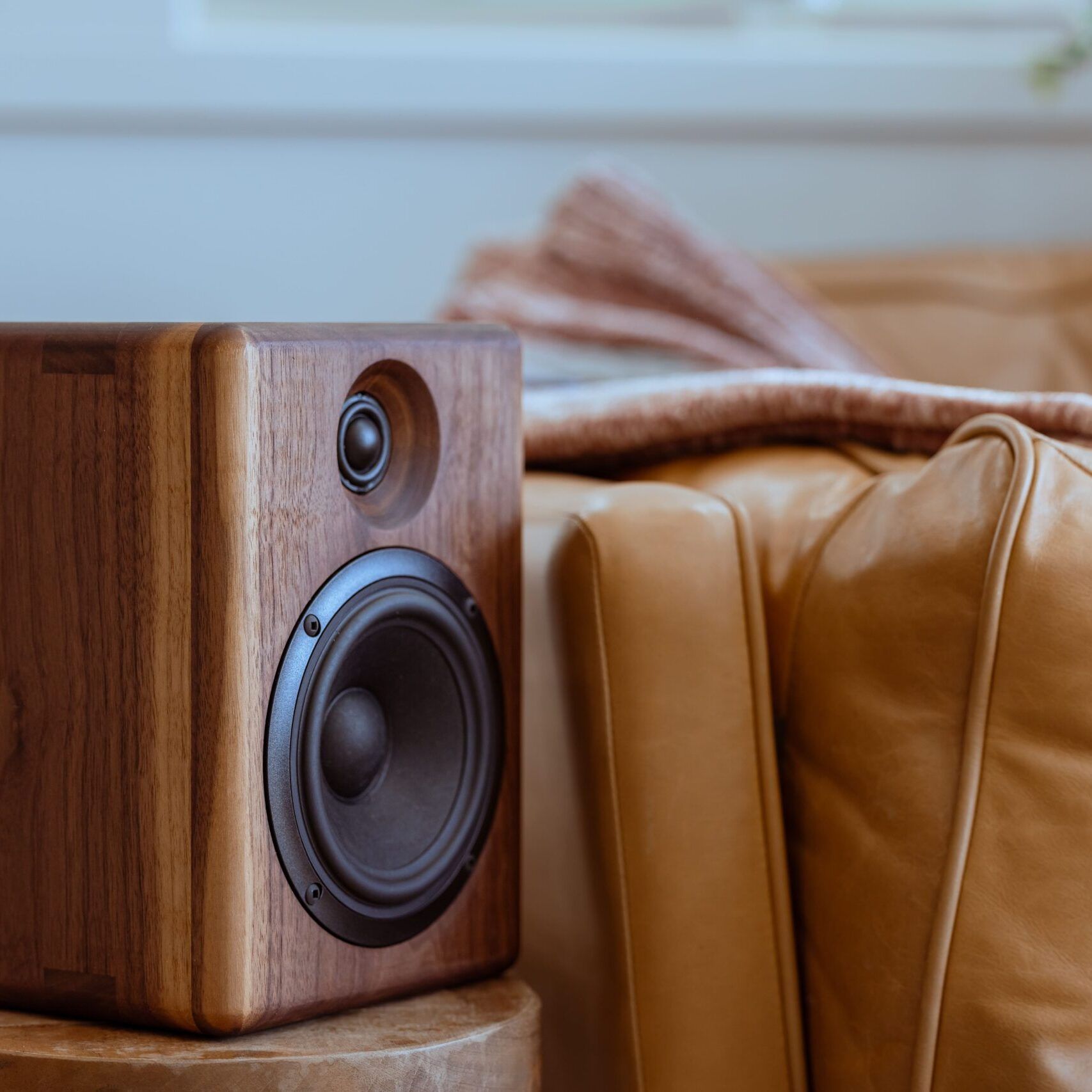 A single Chordwood speaker by Cornaggia, featuring a dark walnut wood cabinet with rounded edges, rests on a small round wooden stool. The speaker is positioned next to a large, cushioned brown leather armchair draped with a light-colored blanket. The shallow depth of field blurs the background, focusing attention entirely on the speaker's beautiful wood grain and black drivers within a cozy, warm, and sophisticated living room setting.