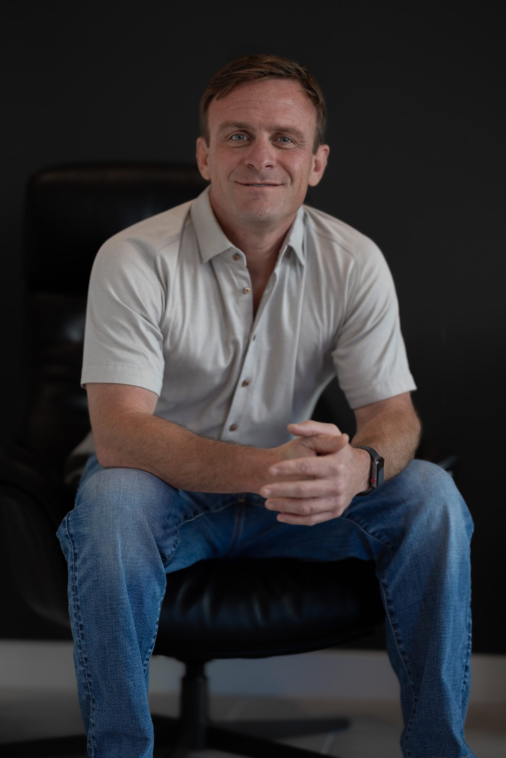 8P0A8931-min A vertical portrait photograph of Luke Cornaggia, a man with short brown hair, sitting in a dark leather office chair. He is wearing a light-colored, short-sleeved button-down shirt and blue jeans. He is looking directly at the camera with a slight smile and his hands are clasped in front of him. The background is a solid, dark black color.